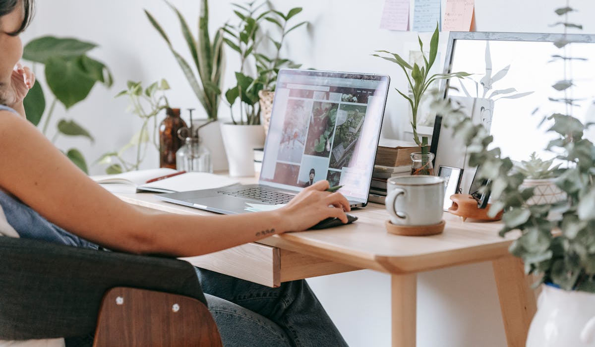 Person focused while working on a laptop at a plant-filled home desk with planner notes on the wall